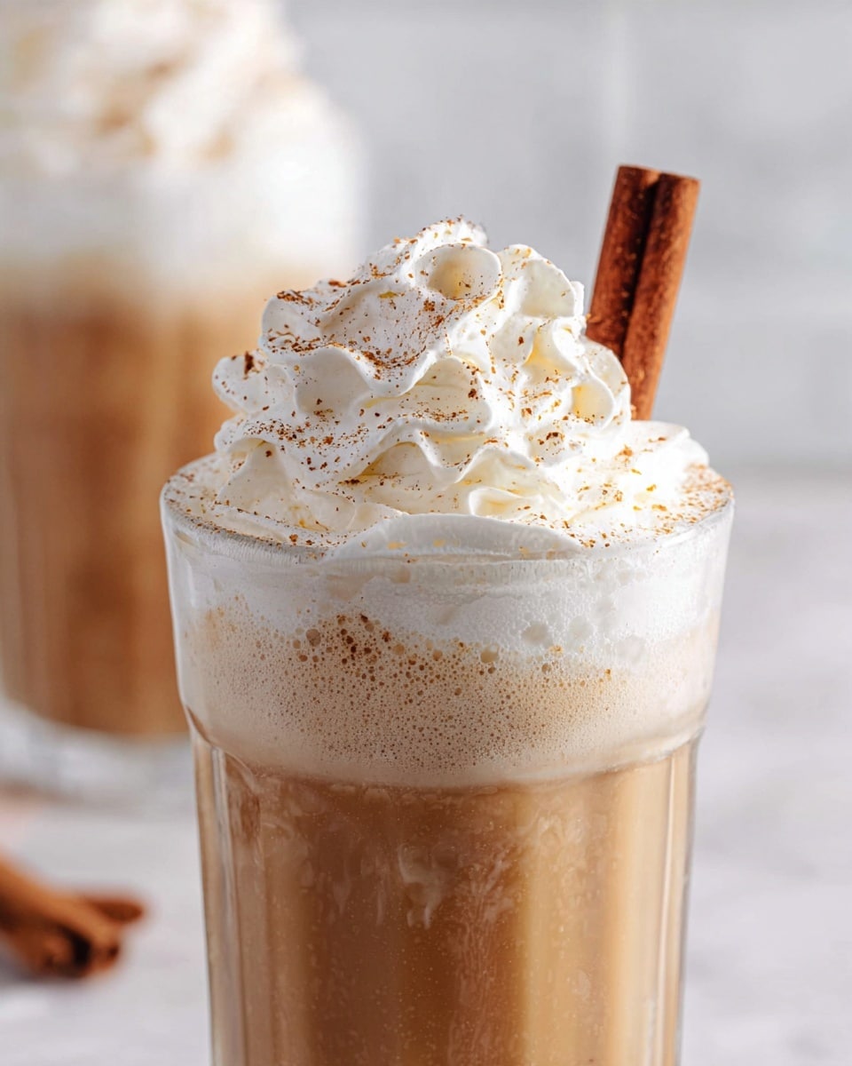 A tall clear glass filled with two layers: the bottom layer is a light brown liquid that looks creamy and smooth, and the top layer is a large swirl of white whipped cream with a soft, fluffy texture. The whipped cream is sprinkled with small brown specks of cinnamon powder and a cinnamon stick is placed upright on the right side of the glass. The glass is set on a white marbled surface with a blurred background showing a second similar glass. Photo taken with an iphone --ar 4:5 --v 7
